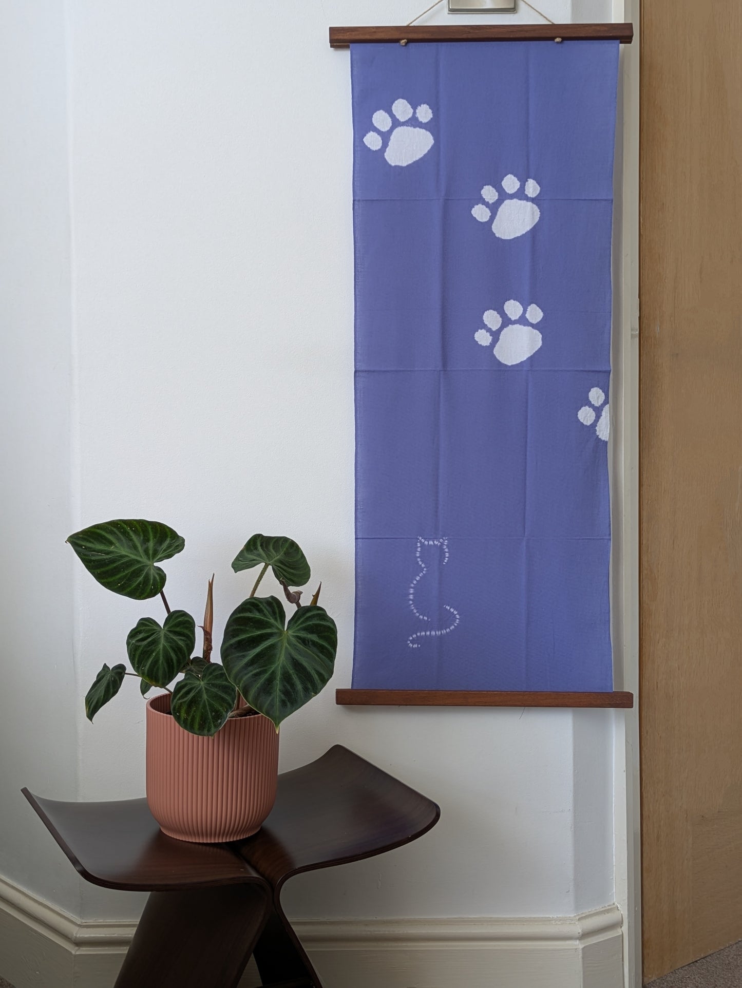 Blue tenugui cloth with white paw prints and a cat silhouette on a wooden rod, with a plant in a pink pot below.