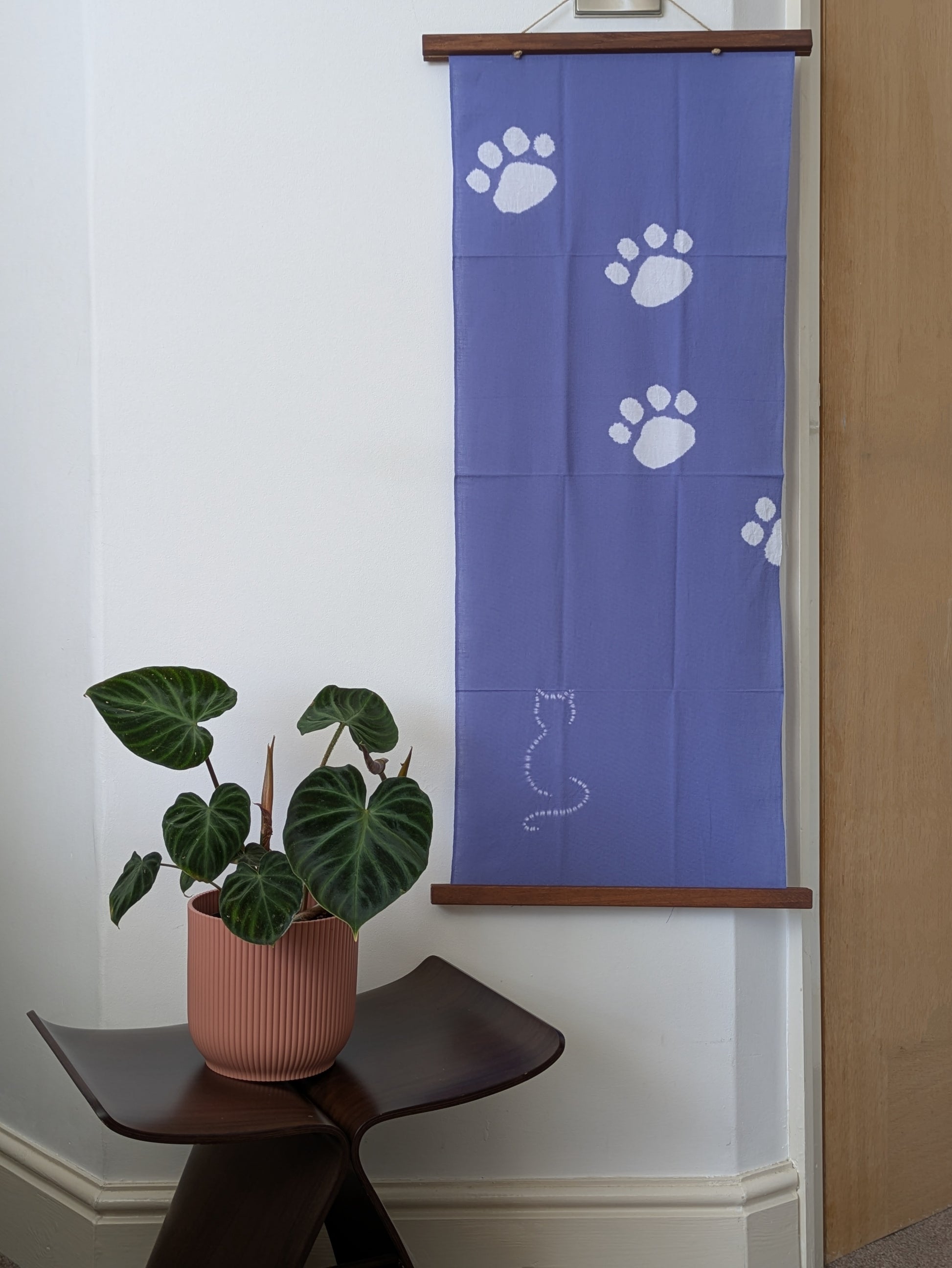 Blue tenugui cloth with white paw prints and a cat silhouette on a wooden rod, with a plant in a pink pot below.