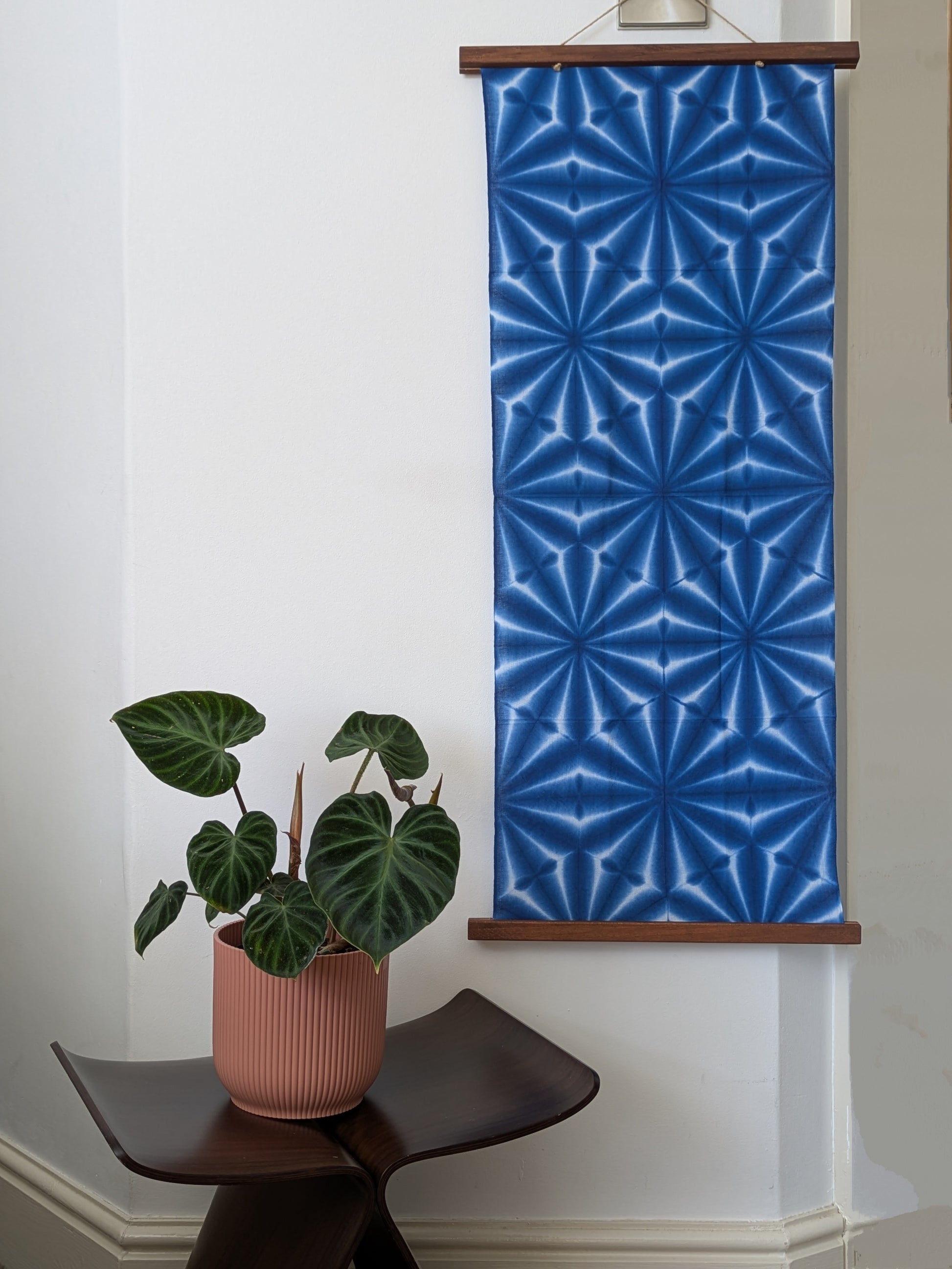 Blue geometric patterned tenugi cloth hanging on a wall next to a potted plant on a table.
