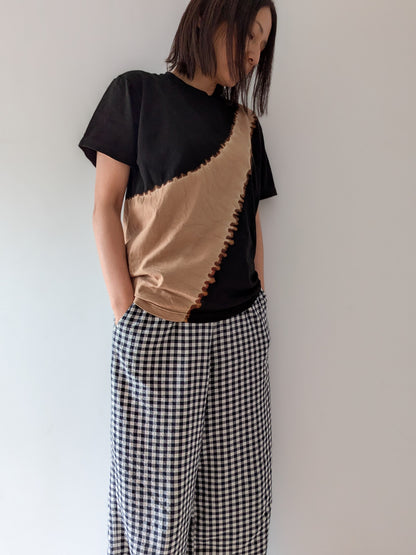A person wearing a black and beige tie-dye t-shirt and black-and-white gingham wide-leg pants, standing against a plain off-white wall.