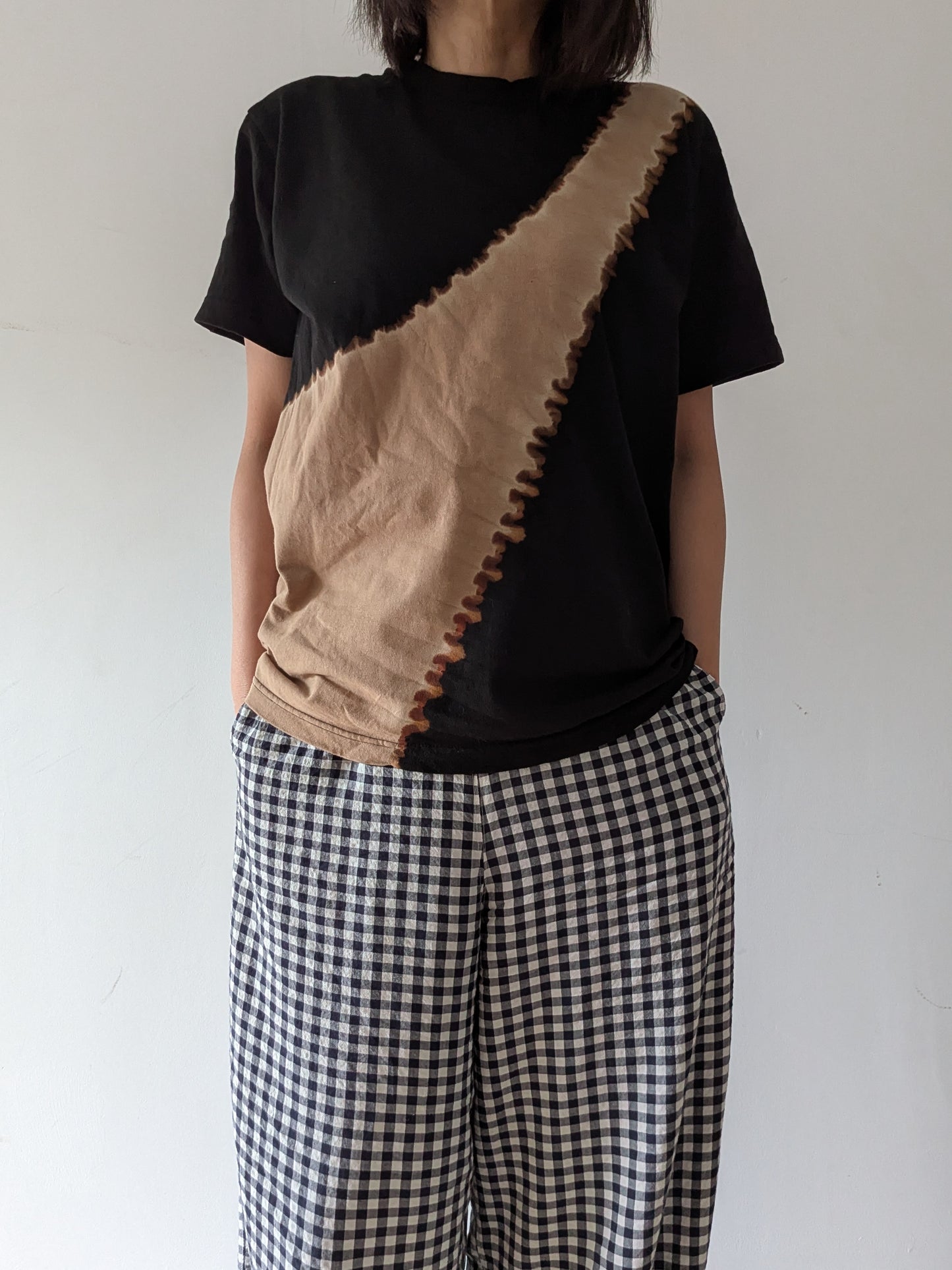 A person wearing a black short-sleeve T-shirt with a large beige diagonal panel and a zigzag stitched seam, paired with black-and-white gingham wide-leg pants, against a plain light background.