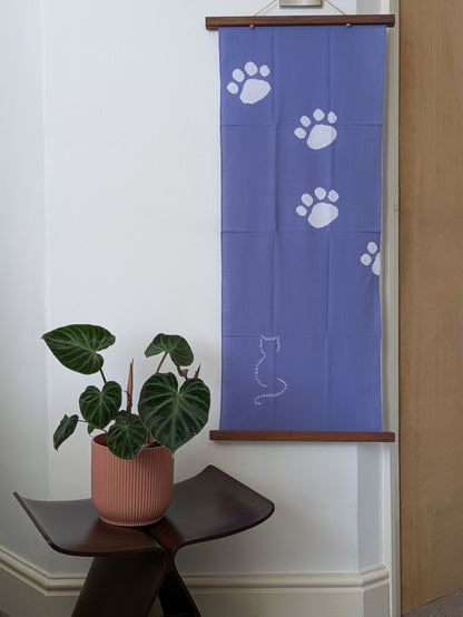Blue tenugui cloth with white paw prints and a cat silhouette on a wooden rod, with a plant in a pink pot below.