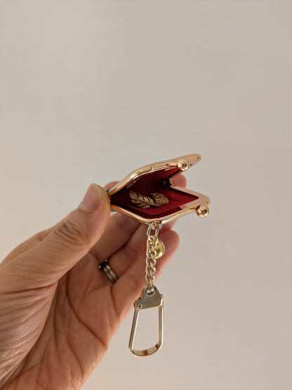 A hand holding a small gold-toned coin purse keychain with a red velvet-lined interior and a gold leaf emblem, against a plain light background.