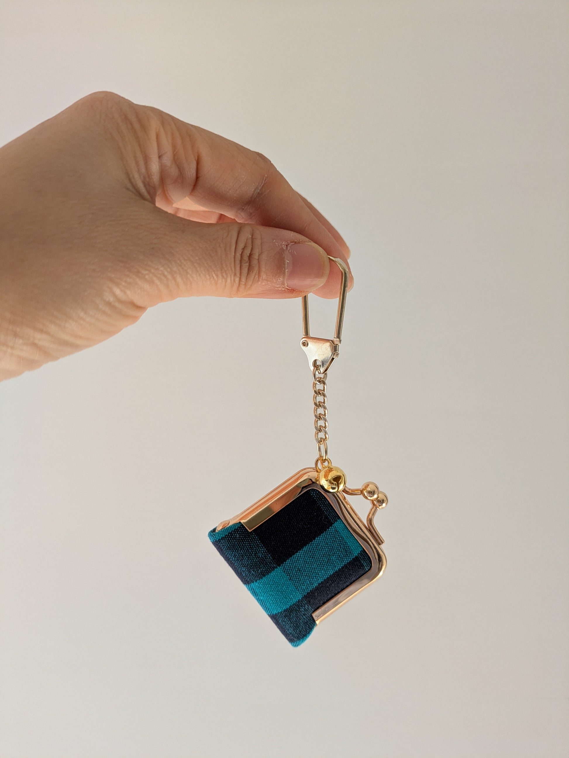 A hand holding a small coin purse keychain with a gold-tone clasp and chain. The coin purse has a striped pattern in teal, dark blue, and black velvet-like fabric.