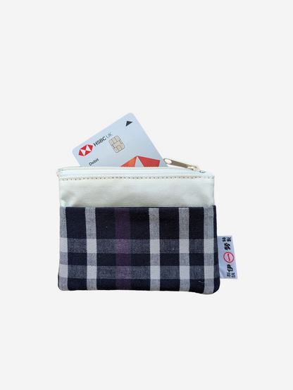 A small zippered pouch with a cream-coloured top section and a navy, gray, and purple plaid patterned bottom section. A credit card is partially inserted into the zipper, and a small fabric tag is visible on the right side.