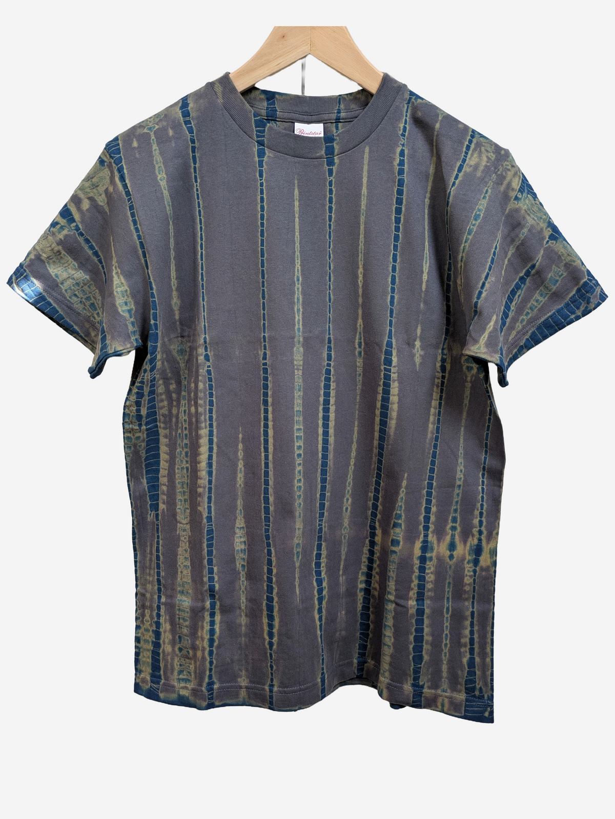 A short-sleeve crewneck t-shirt hand-dyed in Kyoto, Japan with a vertical tie-dye pattern in blue, teal, and beige on a grey base, hanging on a wooden hanger against a light grey background.