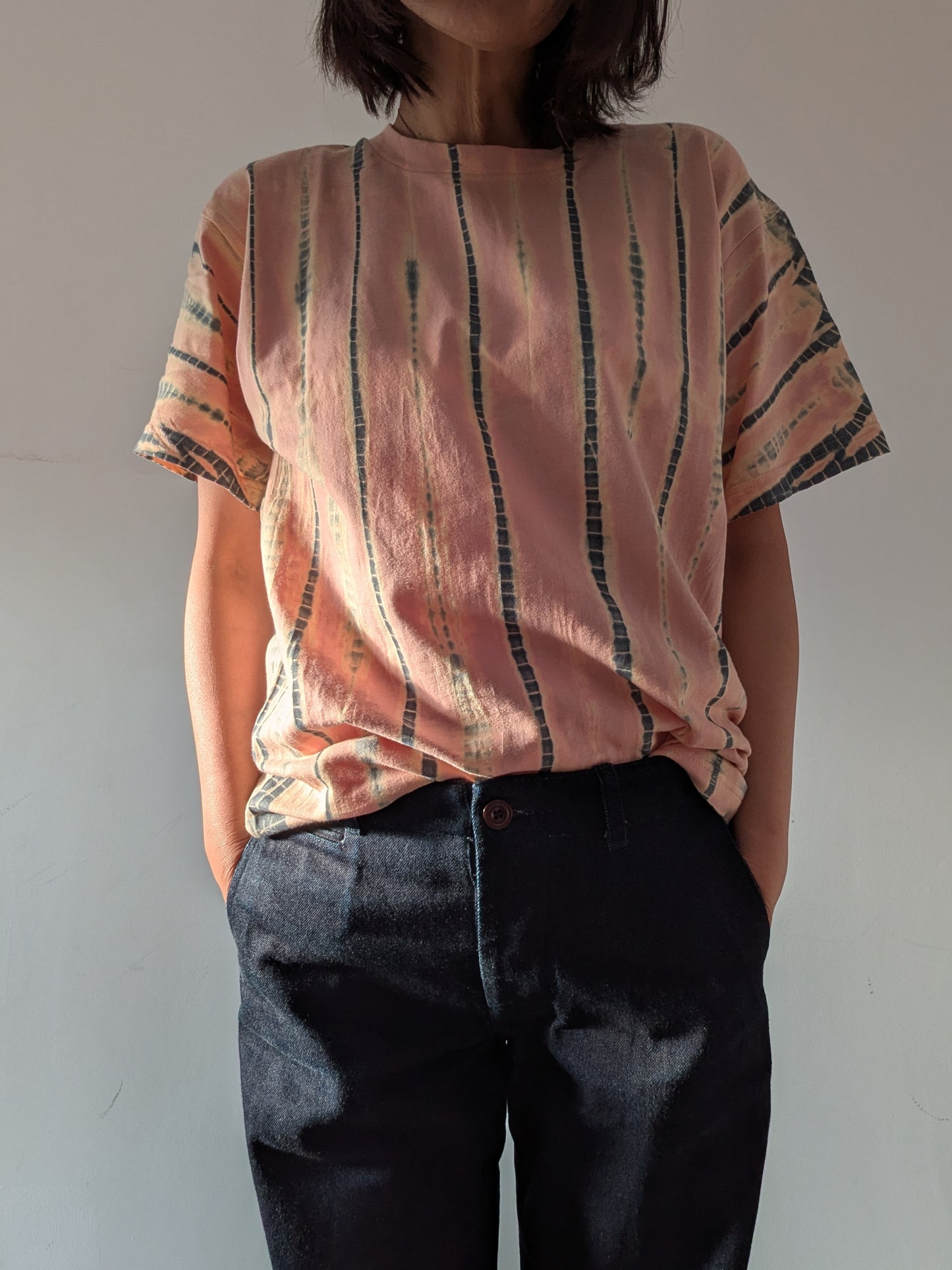 A person wearing a loose short-sleeve top in a dusty peach-pink colour with vertical grey and beige tie-dye stripes, paired with dark denim jeans, standing against a plain light grey wall.