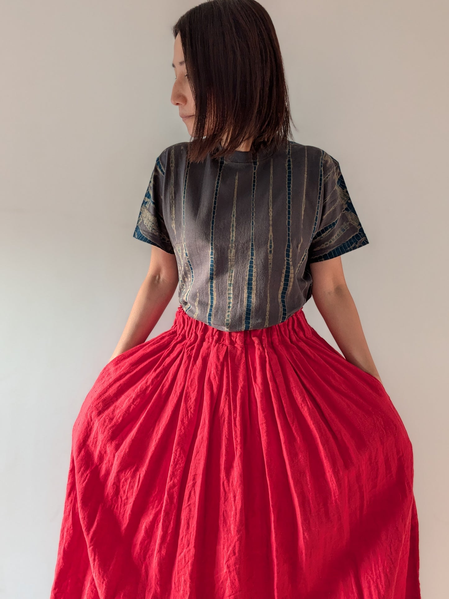 A woman shown wearing a short-sleeve top which hand-dyed in Kyoto, Japan with a dark grey and blue vertical textured pattern and a voluminous bright red skirt, set against a plain light grey wall.