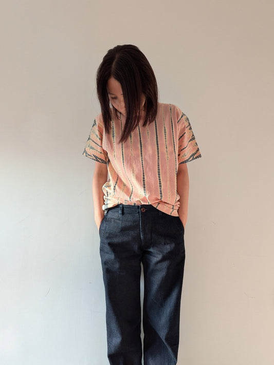 A woman with dark hair stands against a plain light beige wall, wearing a short-sleeve t-shirt with vertical peach, pink, and grey stripes, paired with dark blue jeans.