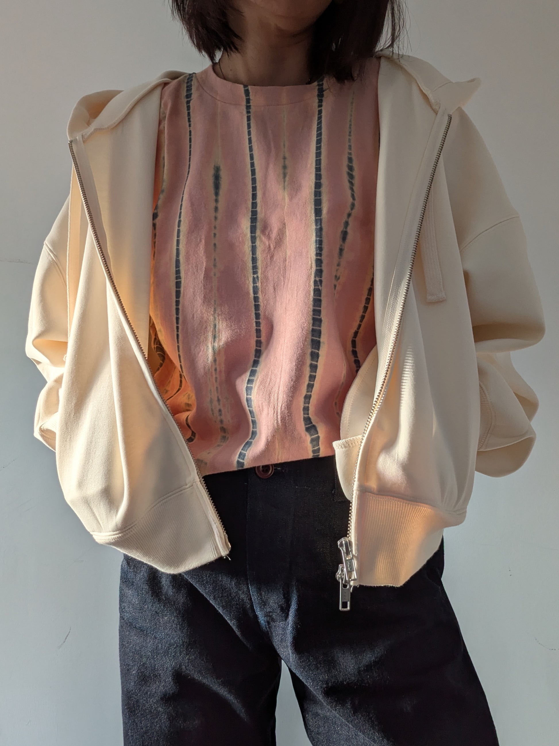 A person wearing a loose short-sleeve top in a dusty peach-pink colour with vertical grey and beige tie-dye stripes and a white hoodie paired with dark denim jeans, standing against a plain light grey wall.