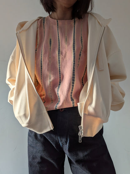 A person wearing a loose short-sleeve top in a dusty peach-pink colour with vertical grey and beige tie-dye stripes and a white hoodie paired with dark denim jeans, standing against a plain light grey wall.