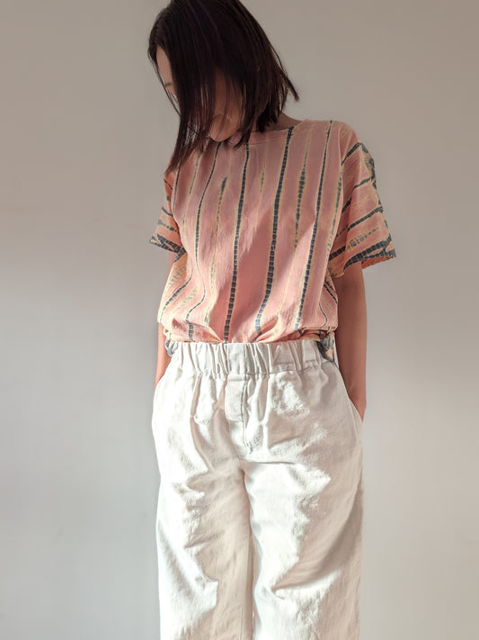 A person wearing a loose short-sleeve top with vertical peach-pink and dark grey stripes, paired with off-white wide-leg pants, standing against a plain light grey wall.