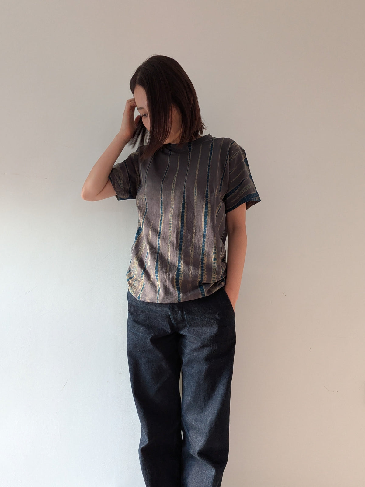 A woman wearing a short-sleeve t-shirt with a vertical striped pattern in grey and blue, paired with dark blue jeans, standing against a plain off-white wall.