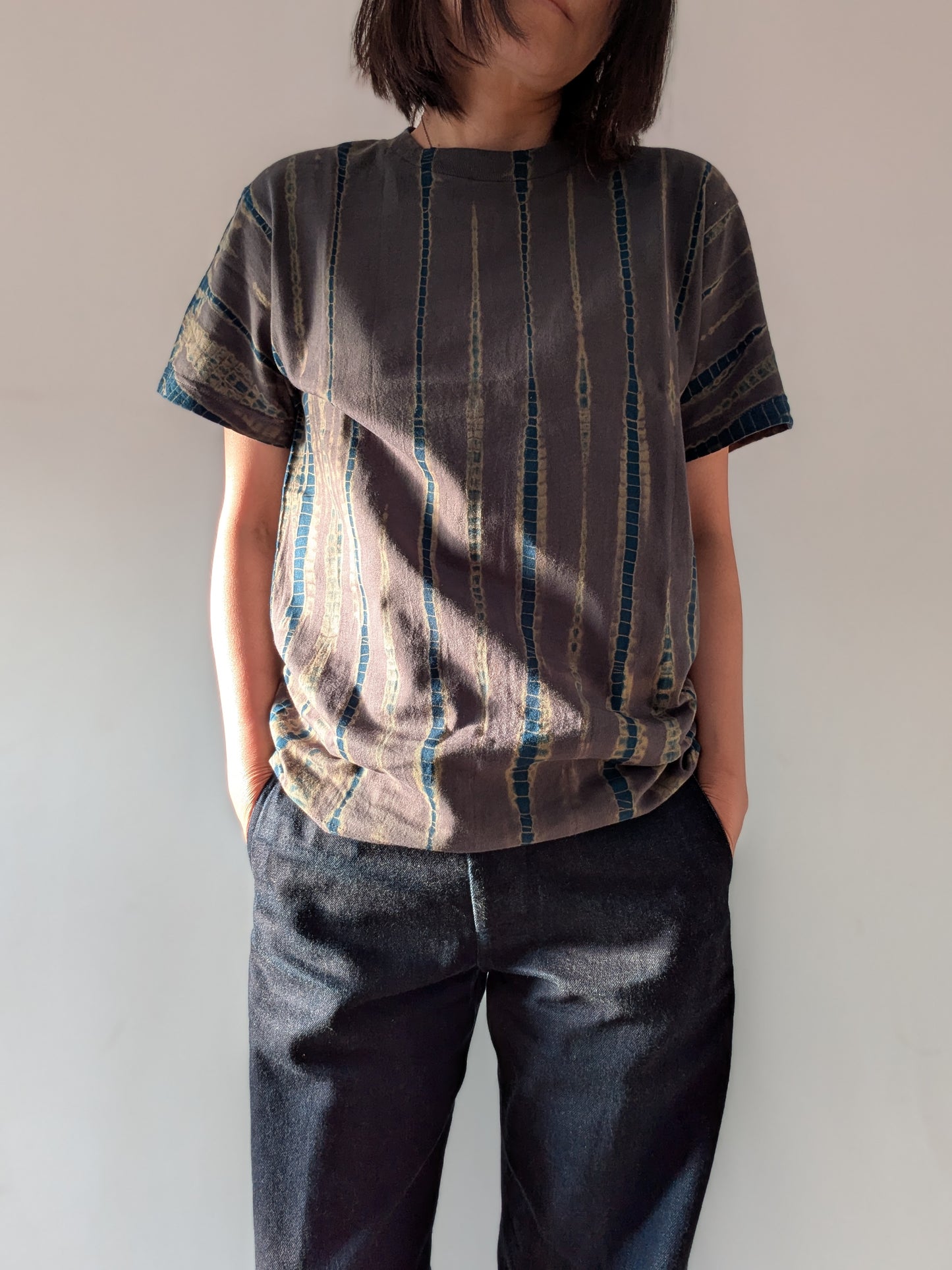 A woman wearing a short-sleeve t-shirt with a vertical striped pattern in grey and blue, paired with dark blue jeans, standing against a plain off-white wall.