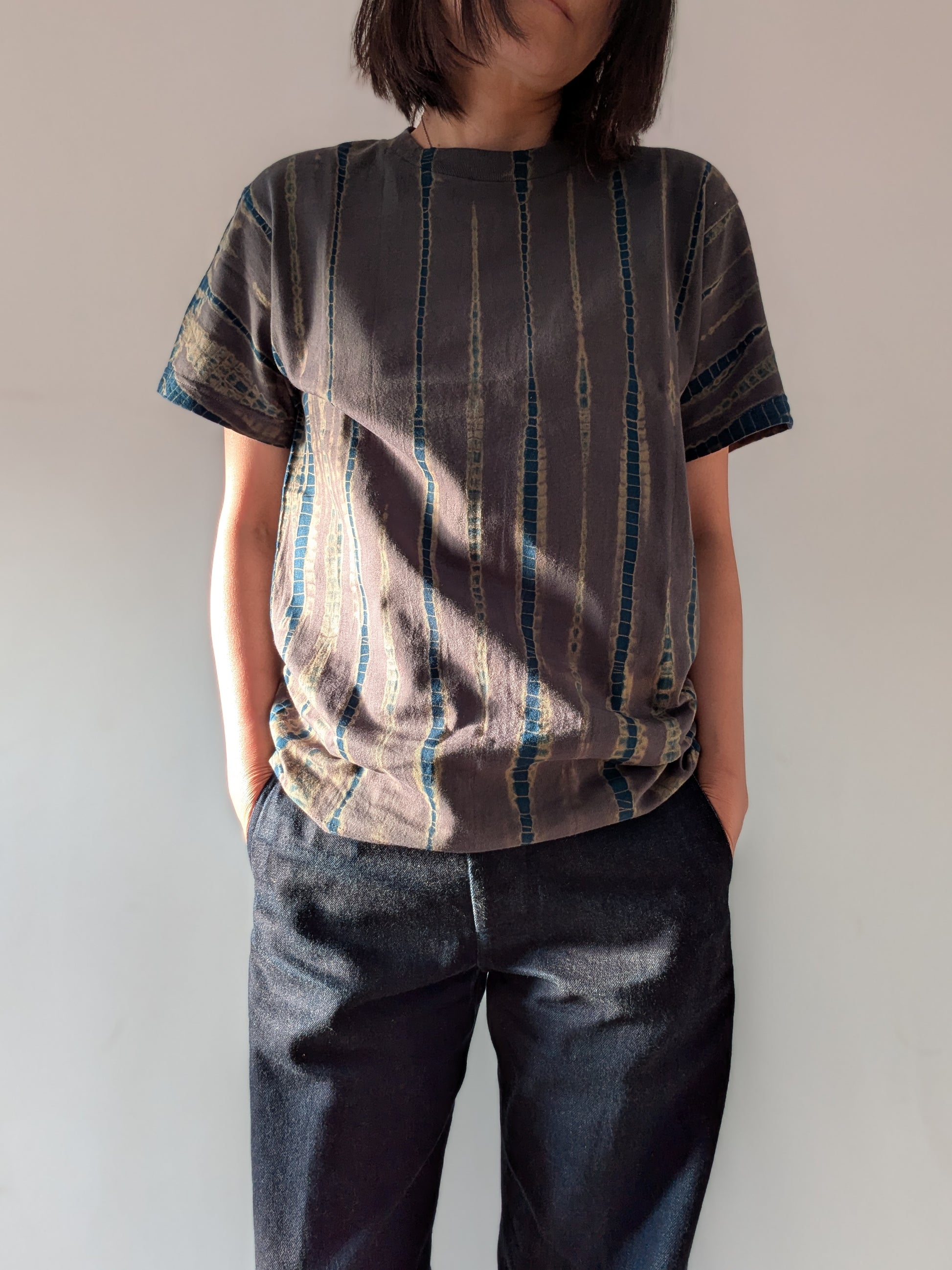 A woman wearing a short-sleeve t-shirt with a vertical striped pattern in grey and blue, paired with dark blue jeans, standing against a plain off-white wall.